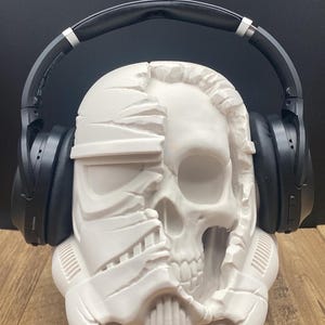 May include: A white skull-shaped headphone stand with a partially exposed Stormtrooper helmet design. Black headphones rest on the stand. The stand is on a wooden surface with a black background.