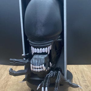 May include: A black Alien Xenomorph figure with a detailed head, sharp teeth, and extended arms and claws. The figure is set within a rectangular frame and sits on a black base. Two business cards are in the foreground.