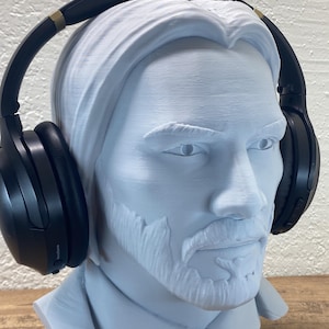 May include: A light blue bust of a man with long hair and a beard, wearing black over-ear headphones. The bust is on a black base, and a black cord extends from the base.
