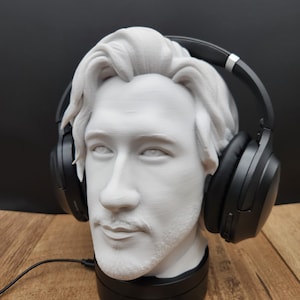 May include: A white bust of a man with headphones resting on his head. The headphones are black. The bust is on a black base. The bust has a mustache and beard. The background is black.