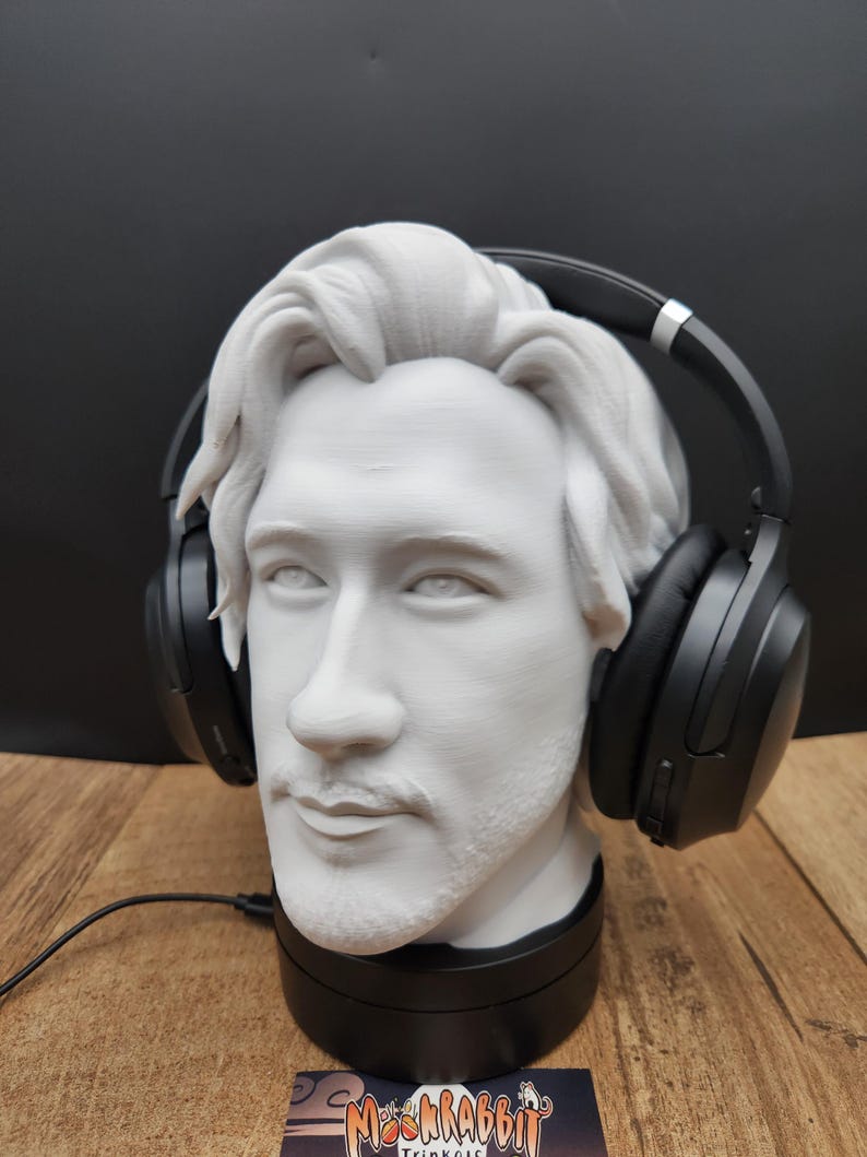 May include: A white bust of a man with headphones resting on his head. The headphones are black. The bust is on a black base. The bust has a mustache and beard. The background is black.