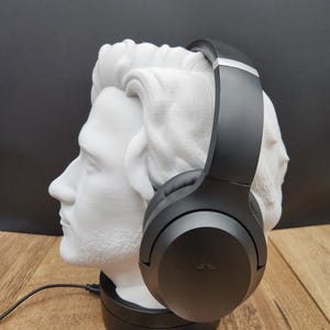 May include: A black pair of headphones rests on a white bust of a man's head. The bust is on a black base, and a black cord extends from the base. The background is black, and the surface is wood-toned.