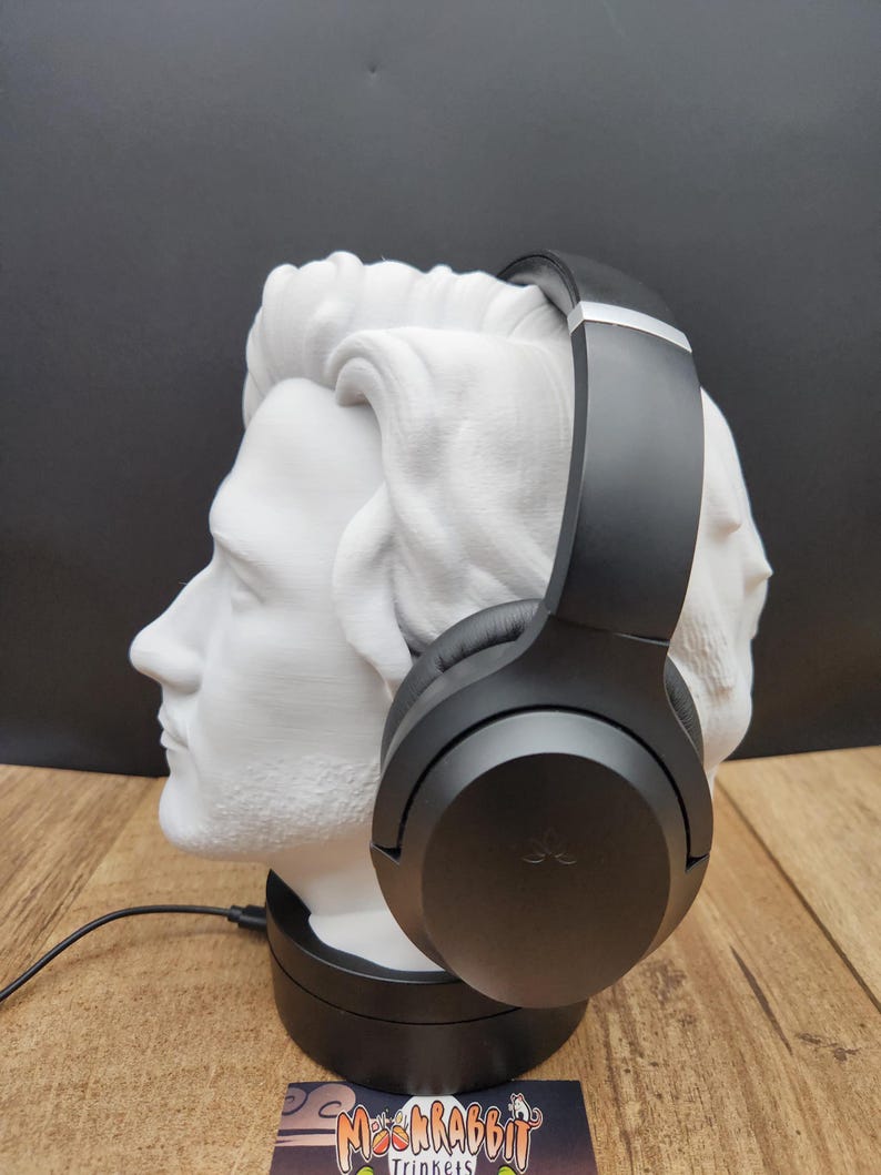 May include: A black pair of headphones rests on a white bust of a man's head. The bust is on a black base, and a black cord extends from the base. The background is black, and the surface is wood-toned.