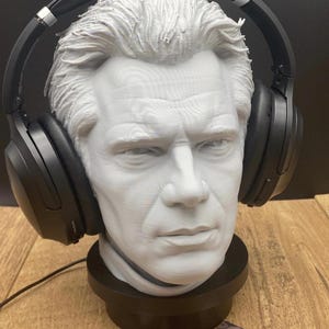 May include: A gray bust of a man wearing black headphones. The bust is mounted on a black base. Two business cards with the "Moonrabbit Trinkets" logo are in front of the bust.