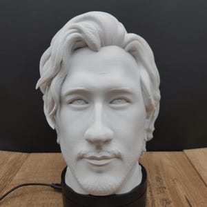 May include: A white bust of a man with light facial hair, mounted on a black rotating base. The bust is detailed with realistic features. A business card with the text "Mschrabbit Trinkets" is visible.
