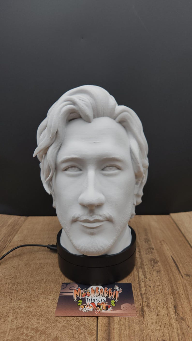 May include: A white bust of a man with light facial hair, mounted on a black rotating base. The bust is detailed with realistic features. A business card with the text "Mschrabbit Trinkets" is visible.