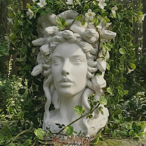 Könnte beinhalten: Eine weiße Medusa-Büste mit Schlangen als Haaren, umgeben von grünem Laub und weißen Blüten. Die Skulptur hat einen ruhigen Ausdruck. Auf dem Sockel der Skulptur steht der Text "MOONRABBIT".