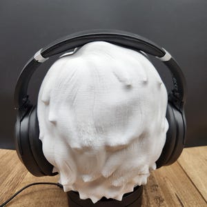 May include: A white, textured head-shaped headphone stand with black headphones resting on top. The stand is on a black base, and a black cable extends from the base. The headphones are black with silver accents.