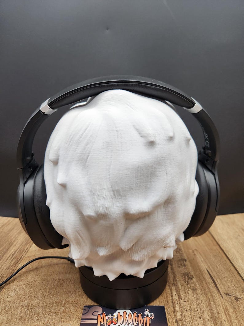 May include: A white, textured head-shaped headphone stand with black headphones resting on top. The stand is on a black base, and a black cable extends from the base. The headphones are black with silver accents.