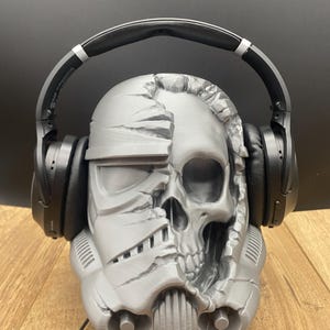 May include: A gray, 3D-printed skull-shaped headphone stand. The design is a fusion of a Stormtrooper helmet and a human skull. Black headphones rest on the stand. The stand is on a wooden surface.