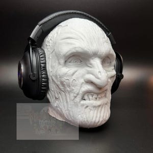 May include: A white, sculpted head with a distressed, aged appearance, wearing black headphones. The head has detailed facial features, including wrinkles and a grimacing expression. The headphones are over-ear style, with black earcups and a padded headband. The overall theme is horror or fantasy.