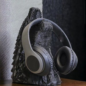 May include: A black Godzilla-shaped headphone stand with a pair of gray and black headphones resting on it. The stand is textured and detailed, with a rough, rocky appearance. The headphones have large, round earcups.