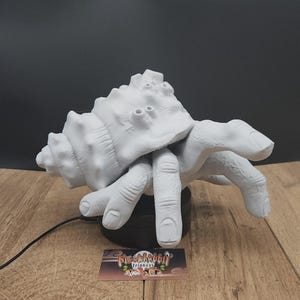 May include: A white sculpture of a hermit crab with a shell and hand-like claws. The sculpture is on a black base, with a business card in front. The background is black and the surface is wood.