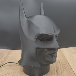 Può includere: Una maschera di Batman nera, con l'iconico design del cappuccio con orecchie a punta e una struttura facciale definita. La maschera è esposta su una base nera, con un biglietto da visita davanti. La maschera è realizzata in materiale opaco.
