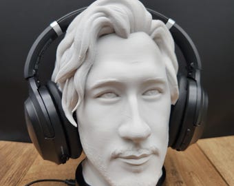 May include: A white 3D-printed bust of a person with wavy hair and facial hair, wearing black over-ear headphones. The bust is mounted on a black circular stand. A small rectangular sign with the text "MOONRABBIT" is visible at the base.