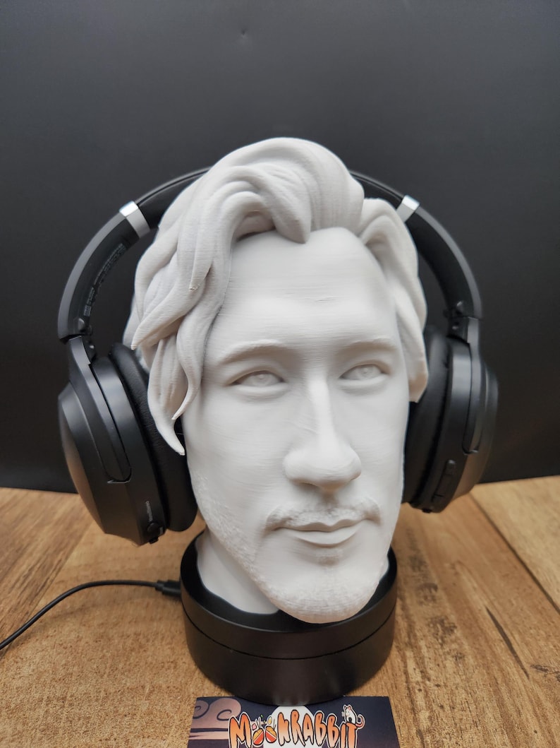 May include: A white bust of a person with headphones resting on the head. The bust is on a black base, and the headphones are black. The bust has a mustache and beard. The background is a dark color.