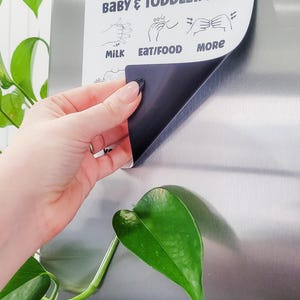 May include: A white magnetic sign with black text and illustrations of hand gestures for baby and toddler sign language. The sign says "Baby & Toddler ASL" and includes the signs for "Milk", "Eat/Food", and "More".