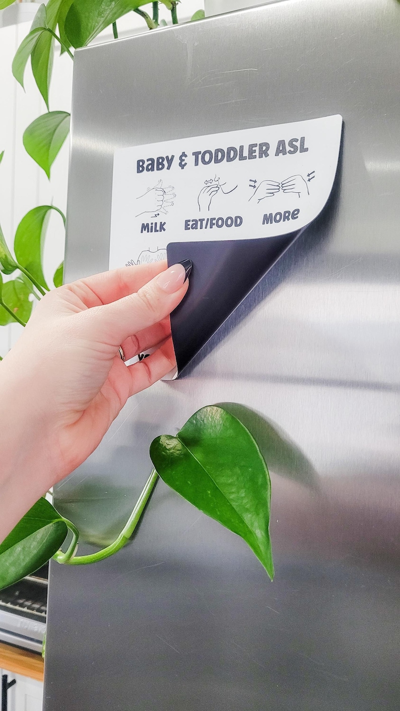 May include: A white magnetic sign with black text and illustrations of hand gestures for baby and toddler sign language. The sign says "Baby & Toddler ASL" and includes the signs for "Milk", "Eat/Food", and "More".