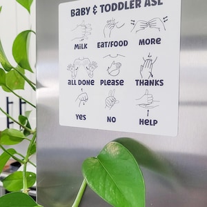 May include: A white magnetic sign with black illustrations of hand signs for baby and toddler ASL. The signs include milk, eat/food, more, all done, please, thanks, yes, no, and help.