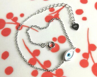 May include: A silver chain bracelet featuring a blue evil eye charm. The charm is set in a silver frame and has a white background. The bracelet has a heart-shaped tag with the number 925 on it.