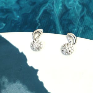 Peut inclure: Une paire de boucles d'oreilles à clous argentées. Chaque boucle d'oreille présente un groupe de petits cristaux transparents de forme ronde, surmontés d'un design incurvé et ouvert. Les boucles d'oreilles sont posées sur une surface blanche sur fond bleu.