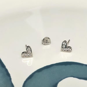 Può includere: Orecchini a bottone a forma di cuore in argento con pietre preziose trasparenti. Gli orecchini sono esposti su una superficie bianca con un design blu e verde. Hanno un perno e un retro.