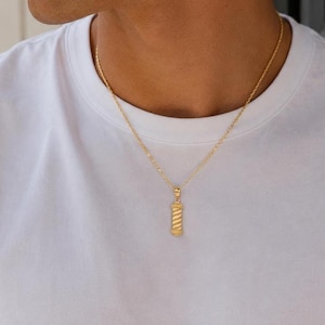 Könnte beinhalten: Eine goldfarbene Halskette mit einer dünnen Kette und einem kleinen, zylindrischen Anhänger mit diagonalem, spiralförmigem Texturmuster. Der Anhänger ähnelt einem traditionellen Barbier-Stangen-Design und liegt auf einem weißen T-Shirt.