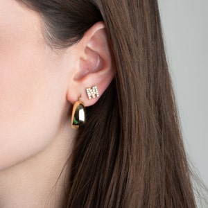May include: A pair of gold earrings. One earring is a simple hoop shape, while the other is a small letter 'M' made of pave diamonds.