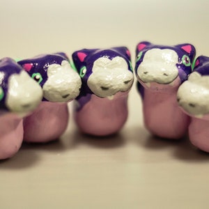 Puede incluir: Cinco pequeñas figuritas pintadas a mano de criaturas moradas y rosas con detalles verdes. Cada figurita tiene una cara blanca con ojos negros y una pequeña boca.
