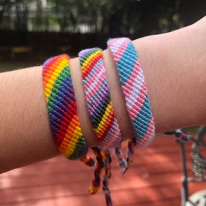 Może przedstawiać: Na nadgarstku widoczne są trzy kolorowe bransoletki przyjaźni. Bransoletki mają wzory tęczy, dumy transseksualist&oacute;w i innych flag dumy. Bransoletki wykonane są z tkanych nici w r&oacute;żnych kolorach, w tym czerwonym, pomarańczowym, ż&oacute;łtym, zielonym, niebieskim i fioletowym.