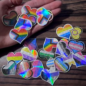 Może przedstawiać: Kolekcja naklejek w kształcie serca z różnymi wzorami flag LGBTQ+. Naklejki zawierają elementy tęczy, holograficzne i wzorzyste. Naklejki znajdują się na drewnianej powierzchni.