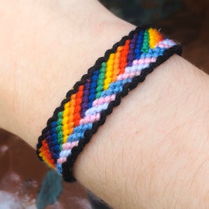 Puede incluir: Un brazalete de amistad arcoíris con hilo negro tejido en el diseño. El brazalete se lleva en la muñeca.