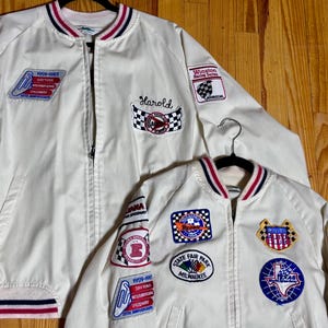 May include: Two cream-coloured vintage racing jackets with various embroidered patches. One jacket features the name "Harold". Patches include "Daytona International Speedway" and "Winston Racing Series". Both jackets have red, white, and blue striped trim.