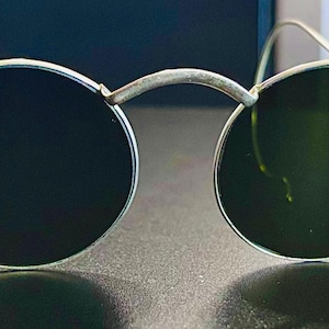 May include: Round sunglasses with dark lenses and a silver-colored metal frame. The glasses have a bridge connecting the two lenses and thin arms that curve around the ears. The lenses appear to be a dark green color.