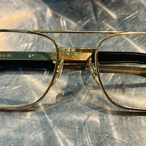 May include: Gold-tone metal eyeglasses featuring a double bridge design and rectangular frames. The inner temple arm is engraved with the text 54/20. The glasses have black temple tips and clear lenses, showcasing a classic aviator-inspired silhouette.