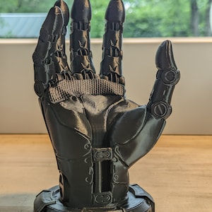 Puede incluir: Una mano robótica negra impresa en 3D con dedos articulados y una base. La mano está en posición con los dedos extendidos.