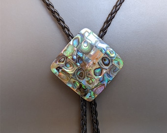 May include: A diamond-shaped bolo tie pendant with iridescent abalone shell inlay. The pendant is attached to a black braided leather cord. The cord has decorative metal tips.