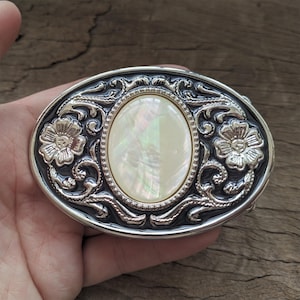 May include: Silver belt buckle with an oval-shaped mother-of-pearl inlay and floral designs.