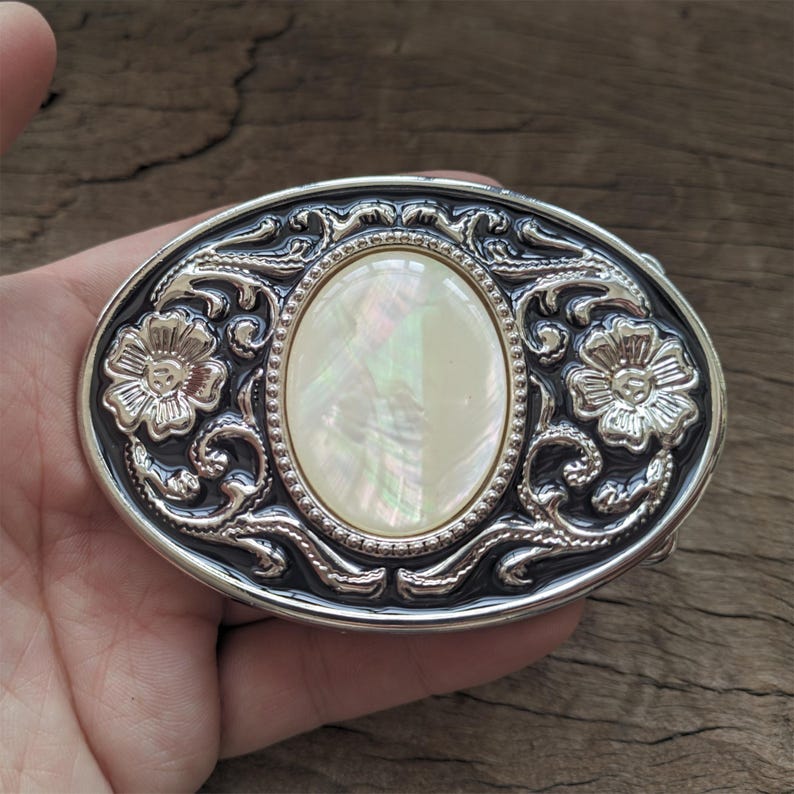 May include: Silver belt buckle with an oval-shaped mother-of-pearl inlay and floral designs.