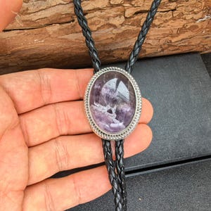 Op de afbeelding: Een bolo-das met een grote, ovale paarse amethist edelsteen in een zilverkleurige metalen zetting met een touwachtige rand. De hanger is bevestigd aan een zwart gevlochten leren koord dat in twee strengen vanaf de centrale zetting naar beneden hangt.