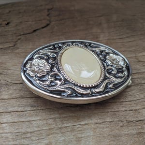 May include: Silver and black oval belt buckle with a white oval stone in the center and floral designs around the stone.