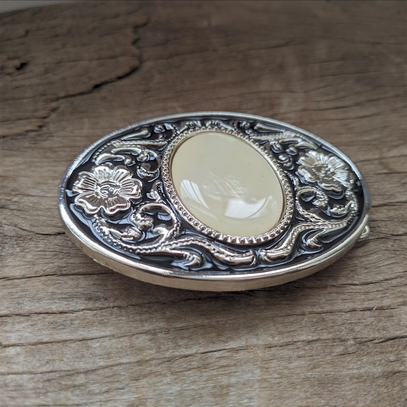 May include: Silver and black oval belt buckle with a white oval stone in the center and floral designs around the stone.
