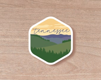 May include: A hexagonal sticker featuring a landscape scene with the word "Tennessee" in cursive. The design includes green hills, a forest, and a yellow and blue sky. The sticker has a white border.
