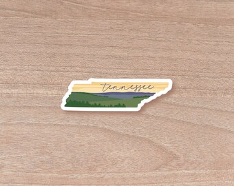 May include: A sticker in the shape of the state of Tennessee. The sticker features a landscape scene with green trees, blue water, and a yellow and tan sky. The word "Tennessee" is written in cursive.