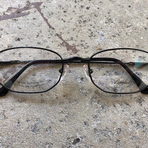 May include: Black oval eyeglasses with the text "Brooks Bros" on the right lens and "Flip-On Sunglasses Available For This Frame" on the left lens. The glasses have a thin black frame.