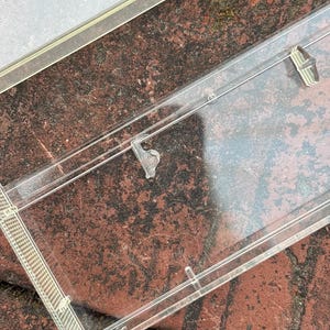 May include: A clear plastic picture frame with a rectangular shape. The frame has a thin, transparent border and small clips to hold a photo in place. The frame is resting on a red brick surface.