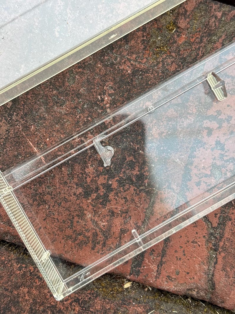 May include: A clear plastic picture frame with a rectangular shape. The frame has a thin, transparent border and small clips to hold a photo in place. The frame is resting on a red brick surface.