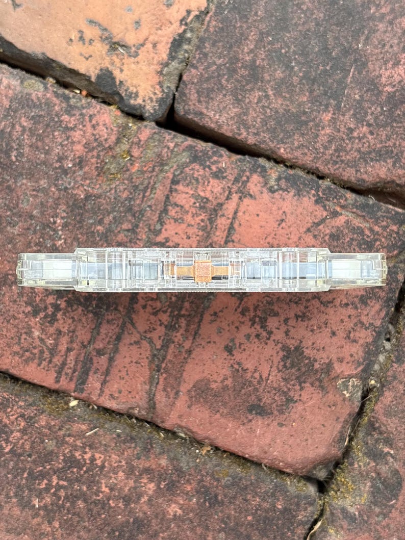 May include: A clear cassette tape with a visible magnetic tape reel. The cassette is resting on a weathered brick surface. The tape's transparent casing allows a view of the internal components, including the tape spools and the magnetic tape itself.