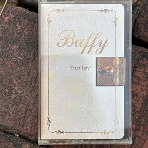 May include: A clear plastic case containing a white cassette tape cover with the word "Buffy" in gold script. The text "FIRST LOVE" is below. A decorative gold clasp is on the right side of the case.
