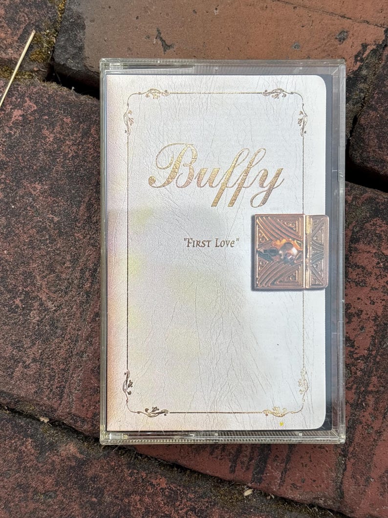 May include: A clear plastic case containing a white cassette tape cover with the word "Buffy" in gold script. The text "FIRST LOVE" is below. A decorative gold clasp is on the right side of the case.